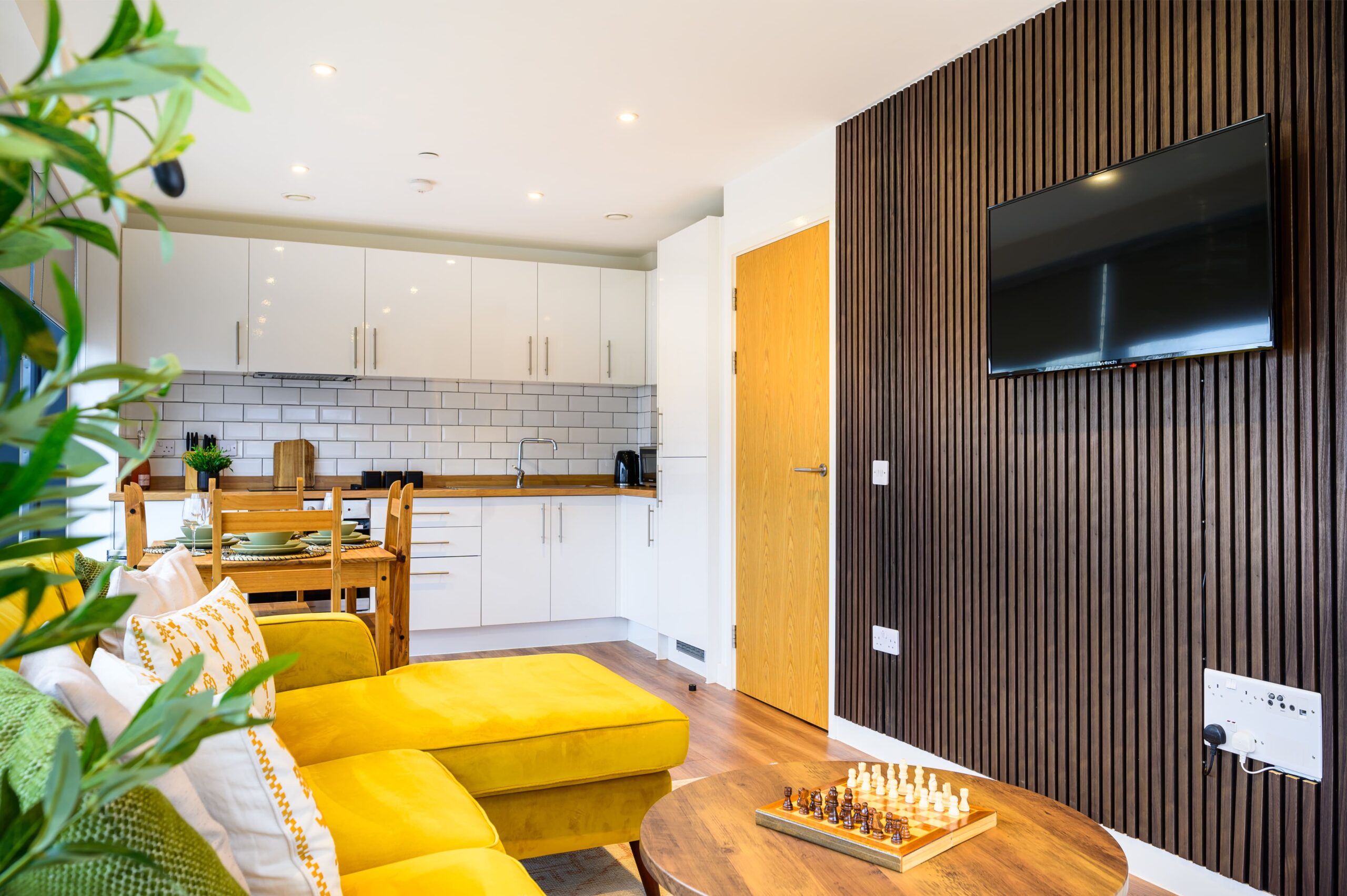 Modern open-plan living area with a yellow sofa, dark wood slatted wall with TV, and white kitchen