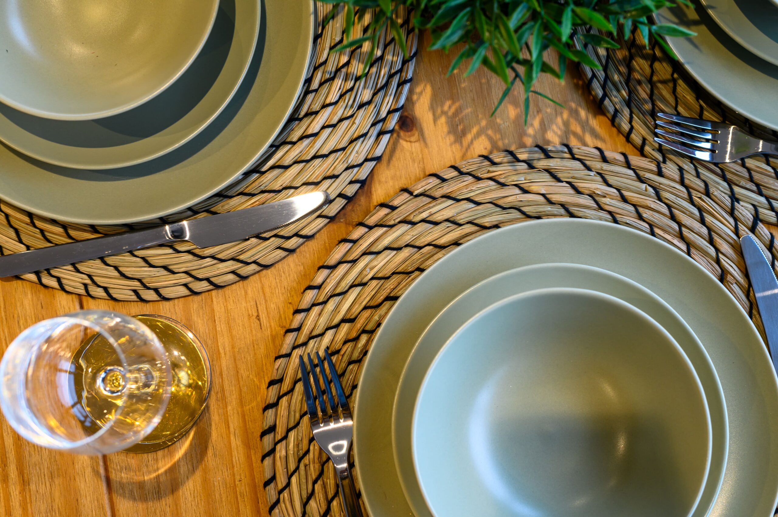 Sage green ceramic dinnerware stacked on woven placemats on a wooden table with silver cutlery and a wine glass
