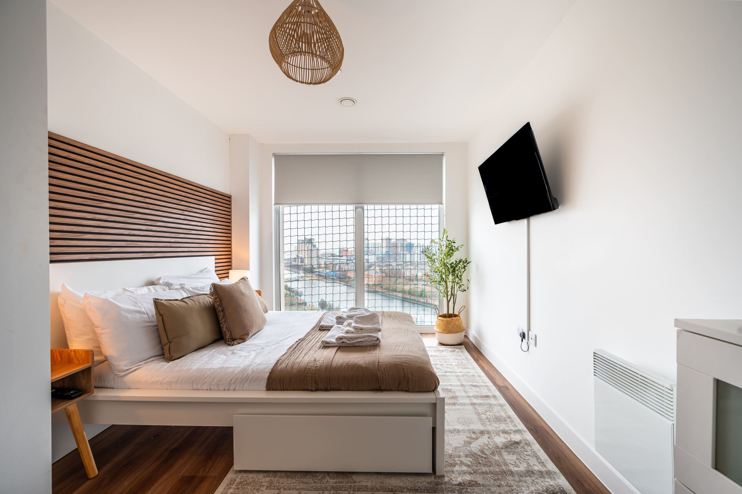 Modern bedroom with a white bed, wooden slat headboard, wall-mounted TV, and a large window with a city and river view