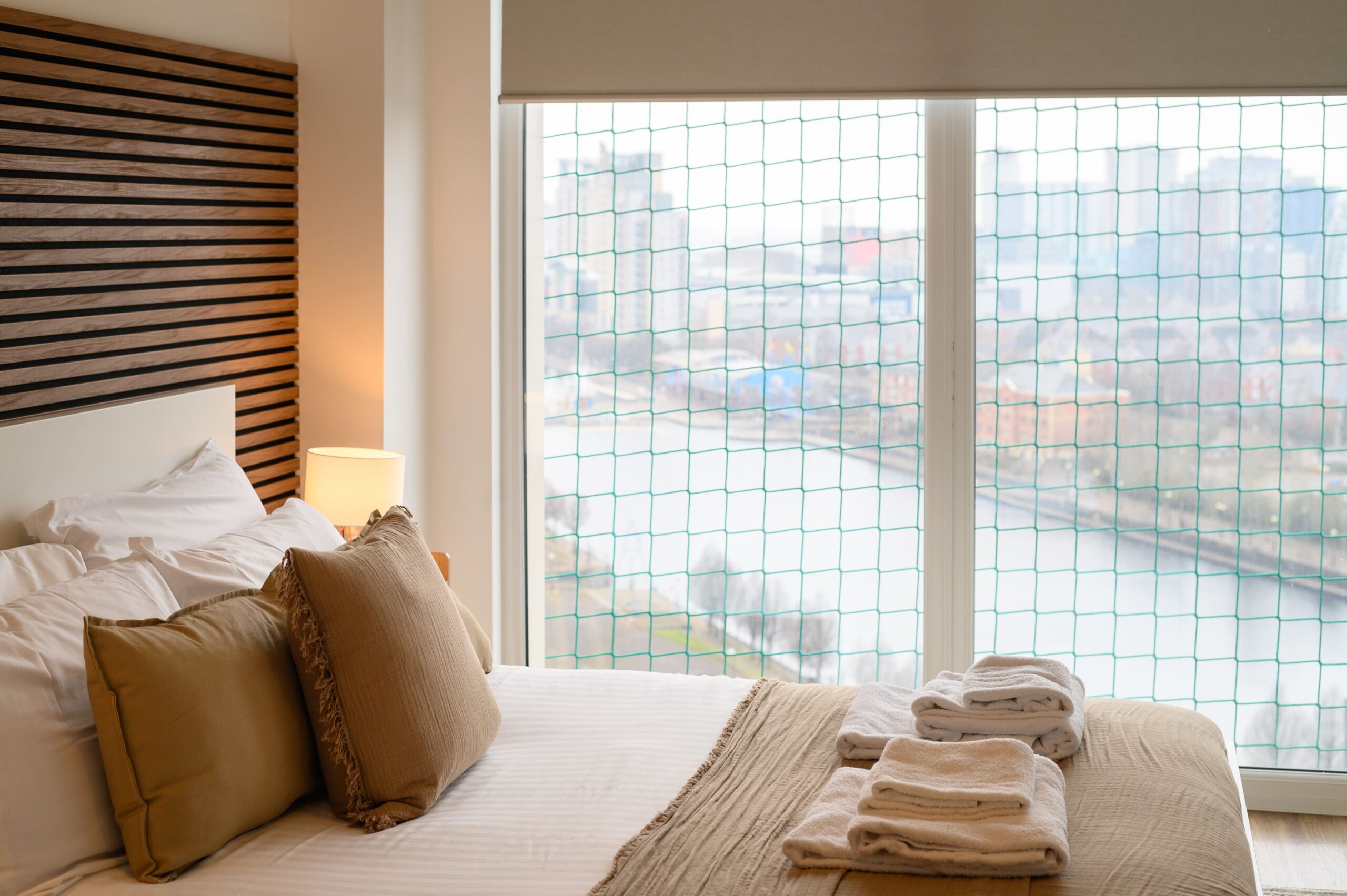 Modern bedroom with a large window overlooking a city waterfront, featuring a bed with beige cushions and towels