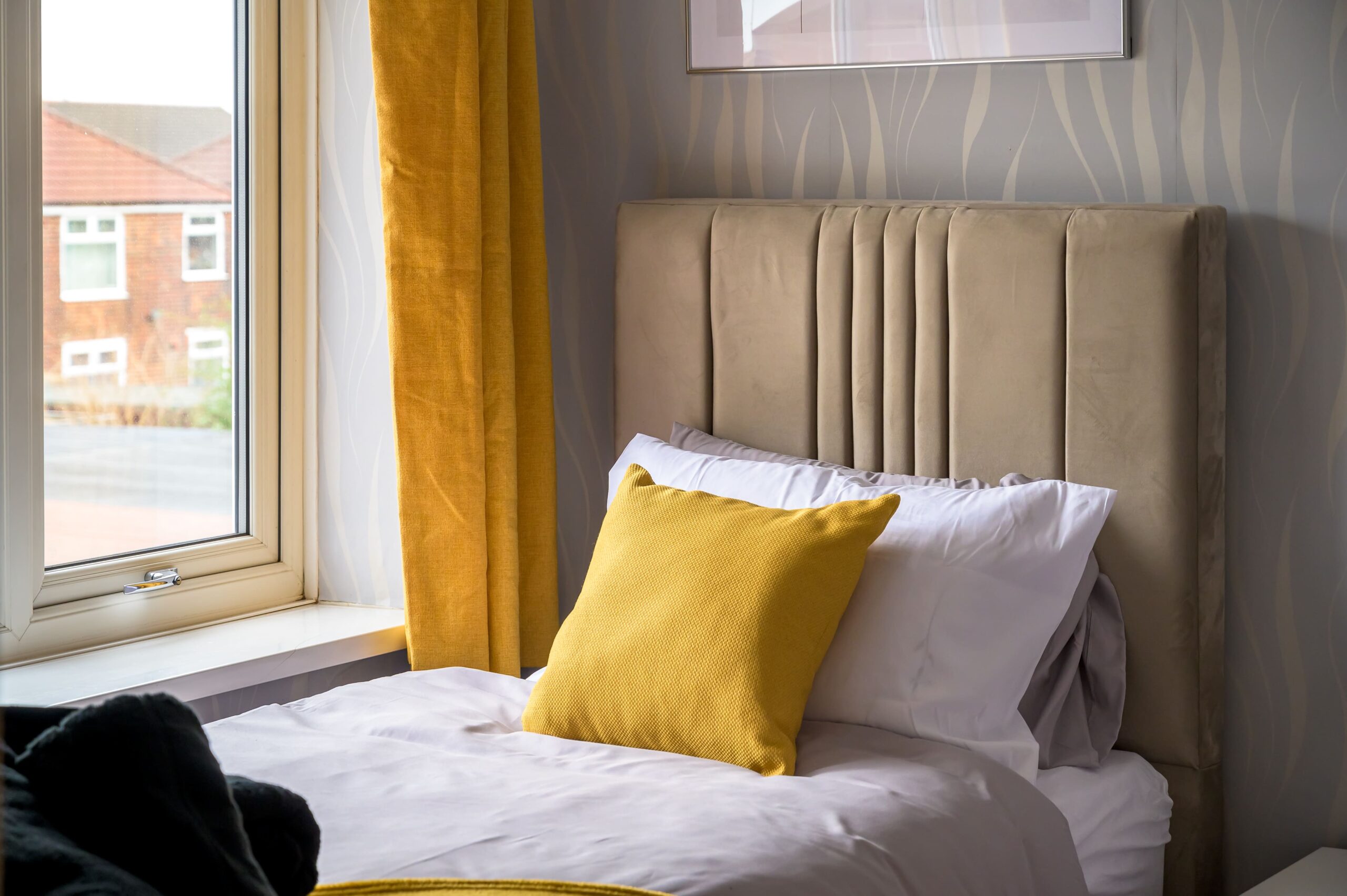 Single bed with a beige tufted headboard, white bedding, and a yellow cushion by a window with yellow curtains