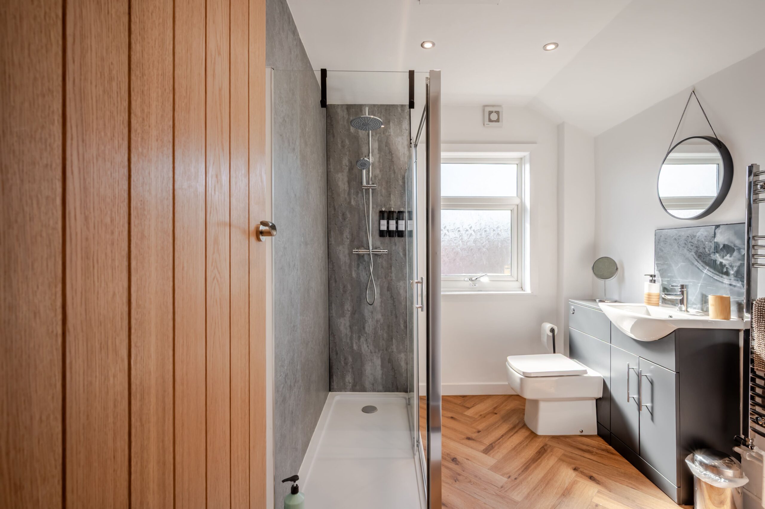 Modern bathroom with walk-in shower, grey vanity unit, white toilet, and herringbone wood flooring