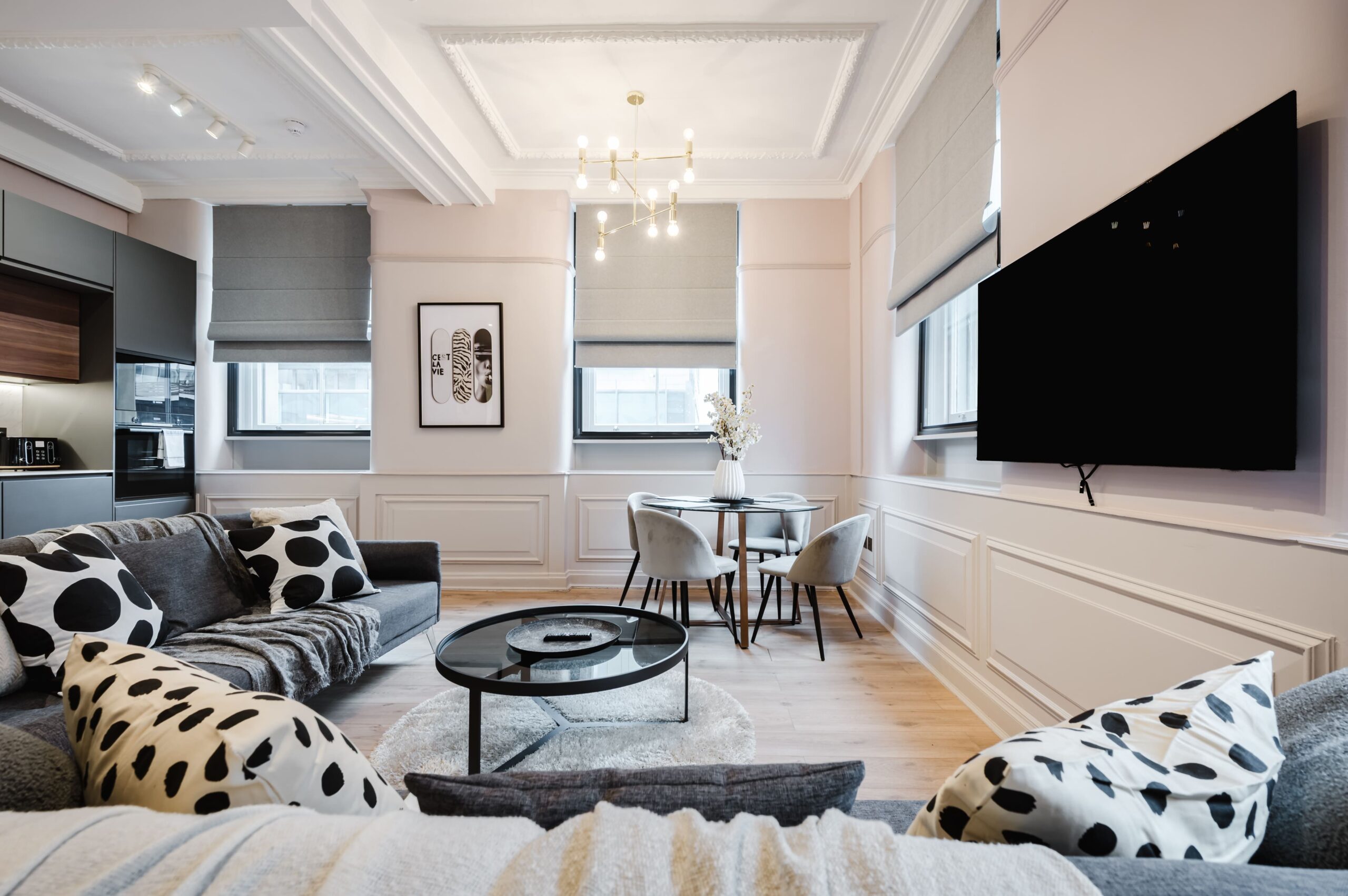 Modern living room with a grey sofa, patterned cushions, glass coffee table, and a small dining area