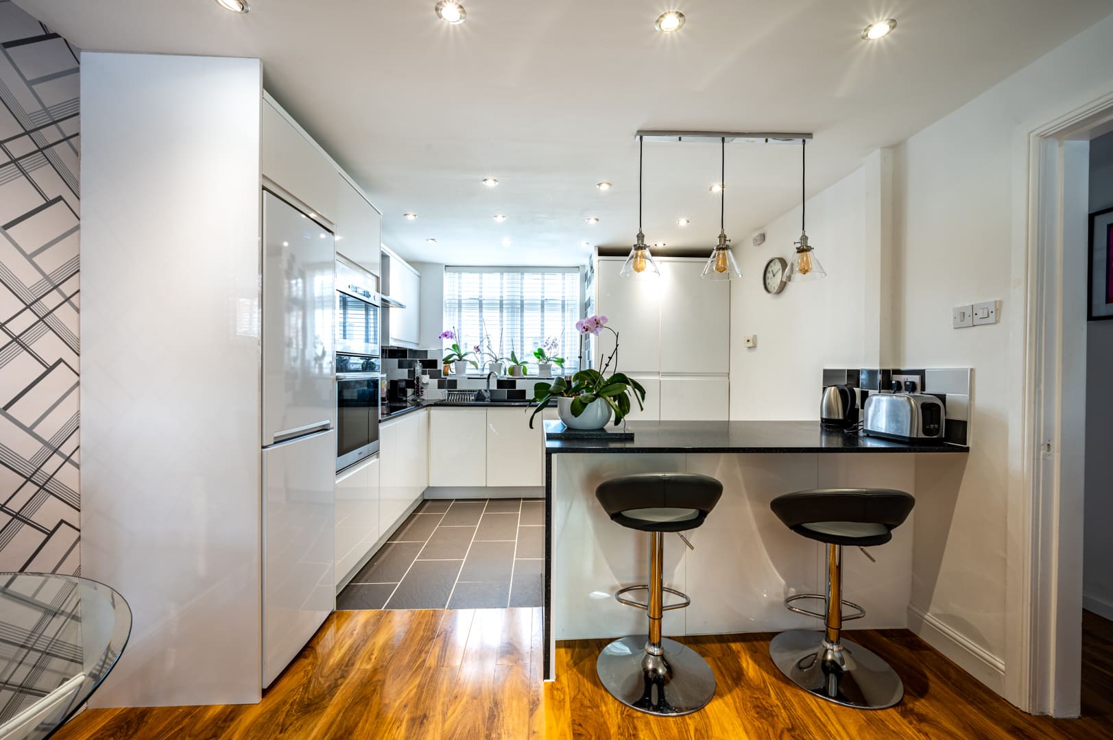 Modern kitchen with white gloss cabinets, black worktops, a breakfast bar with two stools, and integrated appliances