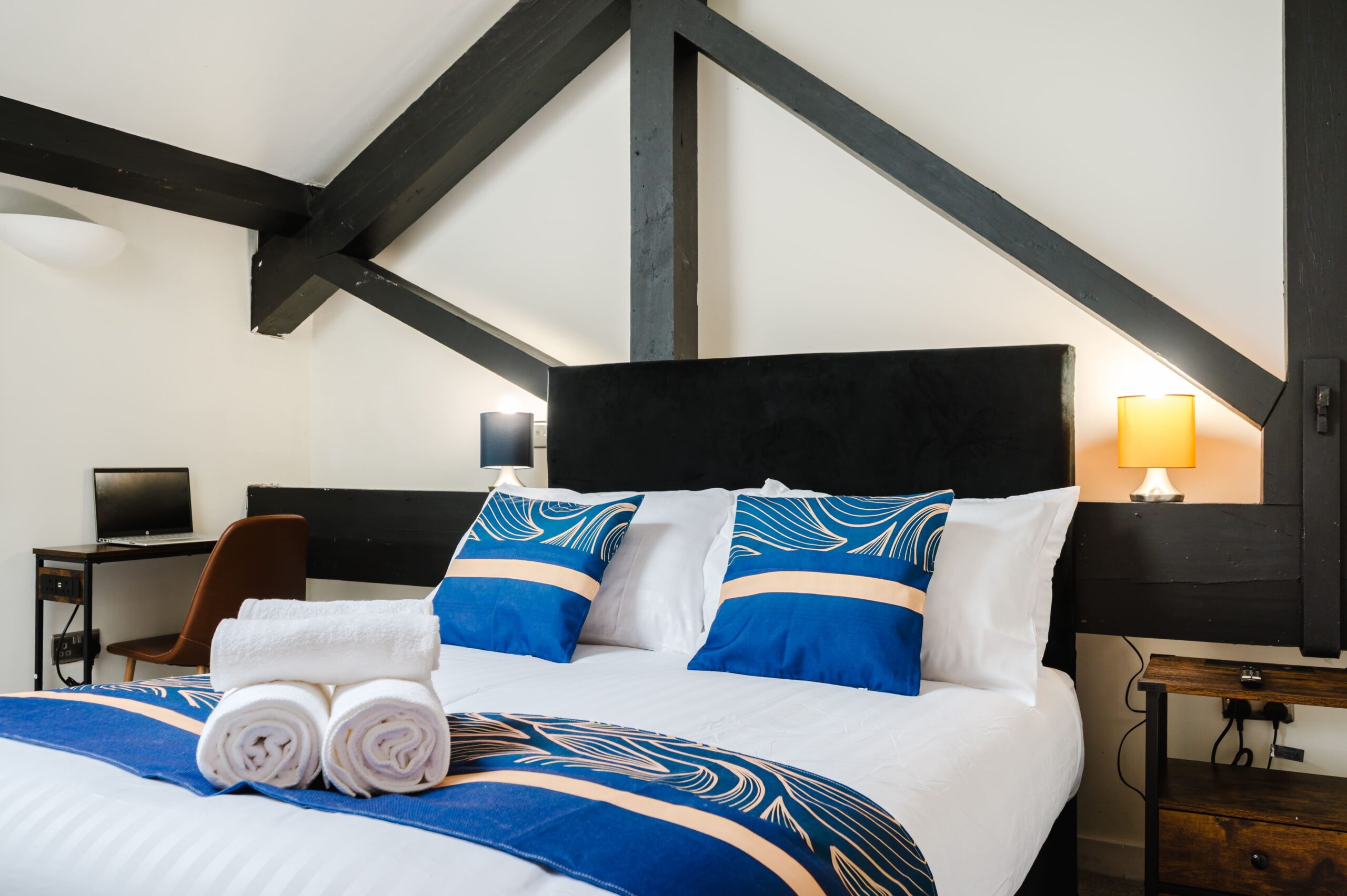 Double bed with blue and gold cushions, white towels, and a black headboard under exposed timber beams