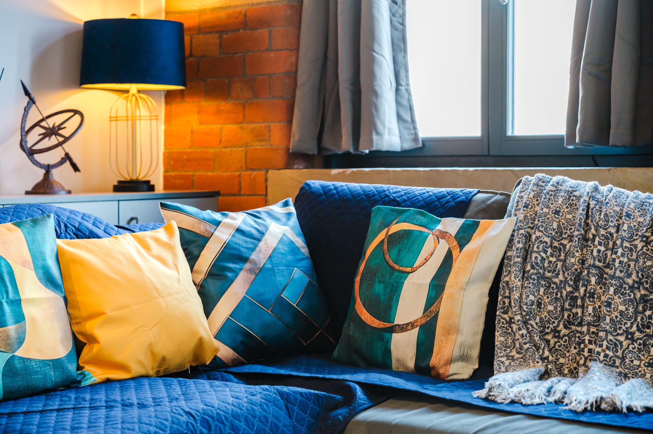 Blue sofa with yellow and patterned cushions, a throw blanket, and a lamp against a brick wall