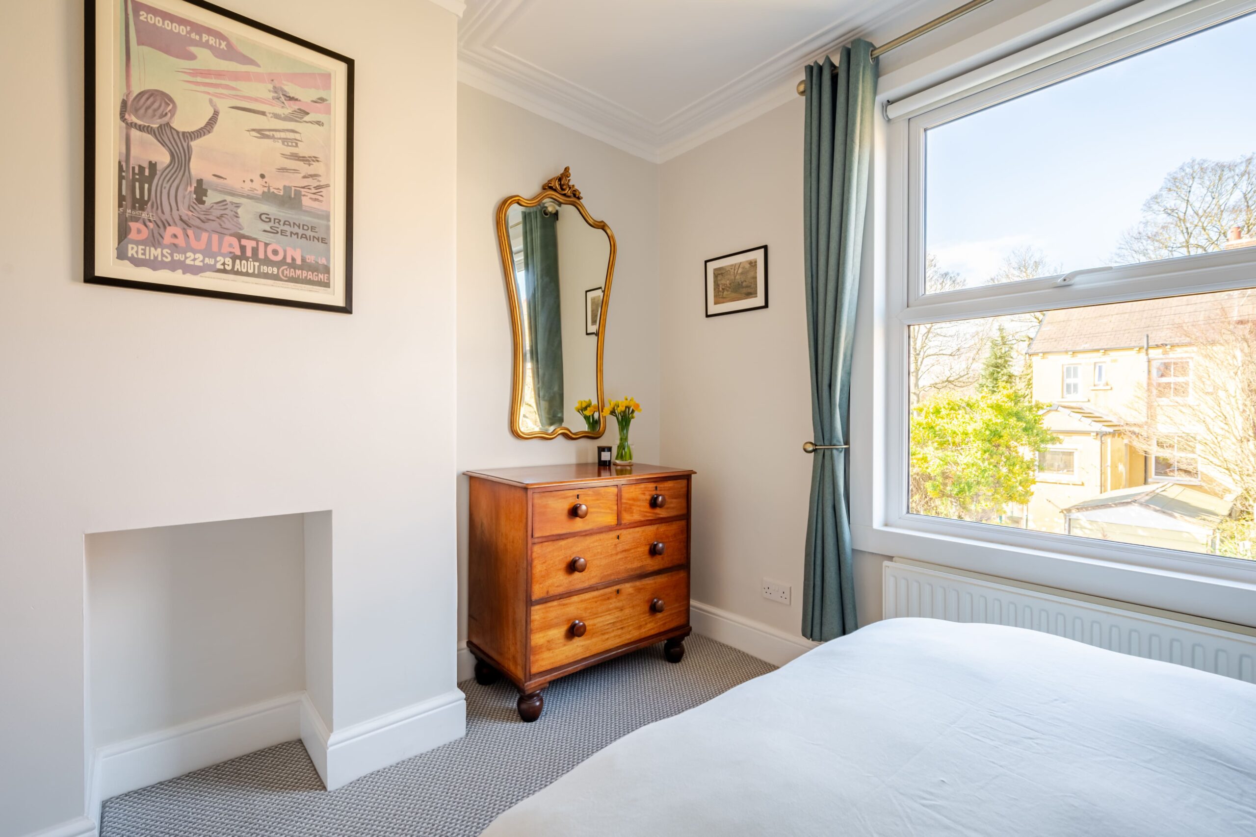 Bedroom featuring a wooden chest of drawers, gold-framed mirror, vintage poster, and window with blue curtains