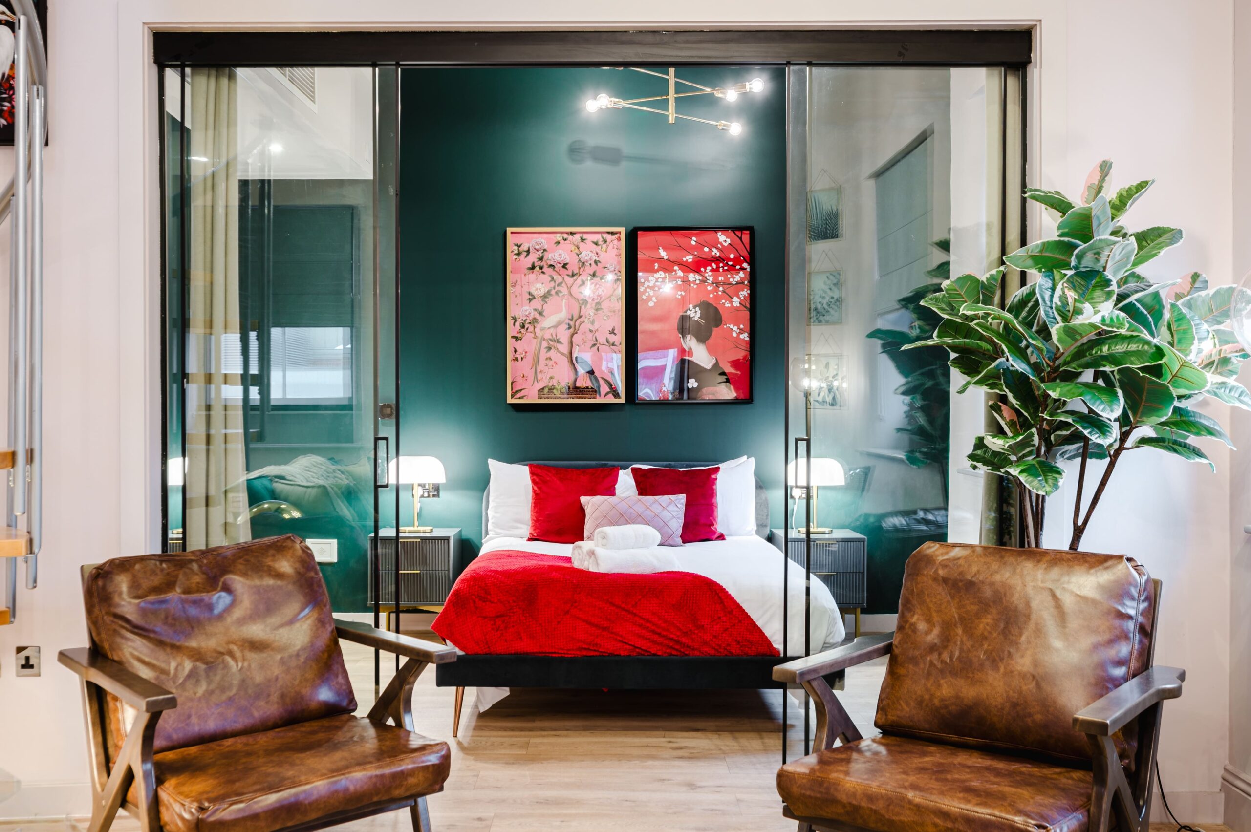 Bedroom with red and white bedding and dark green wall, viewed through glass doors from a living area with leather chairs
