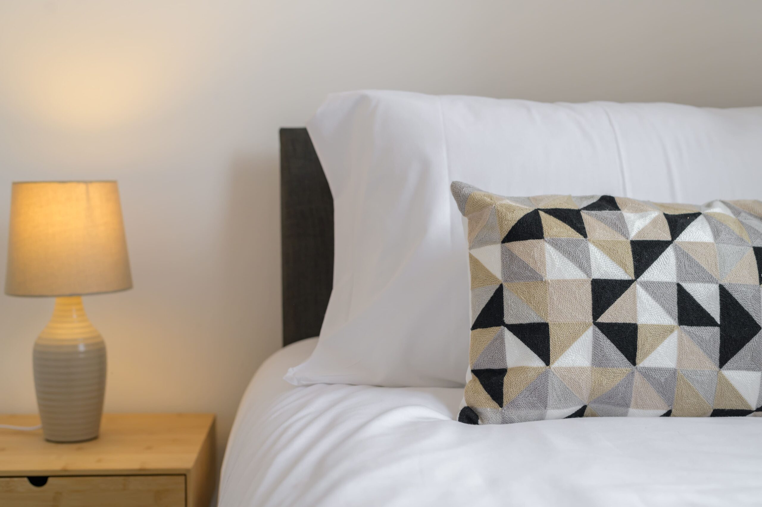 White bed linens with a grey, black, and beige geometric cushion next to a lit bedside lamp