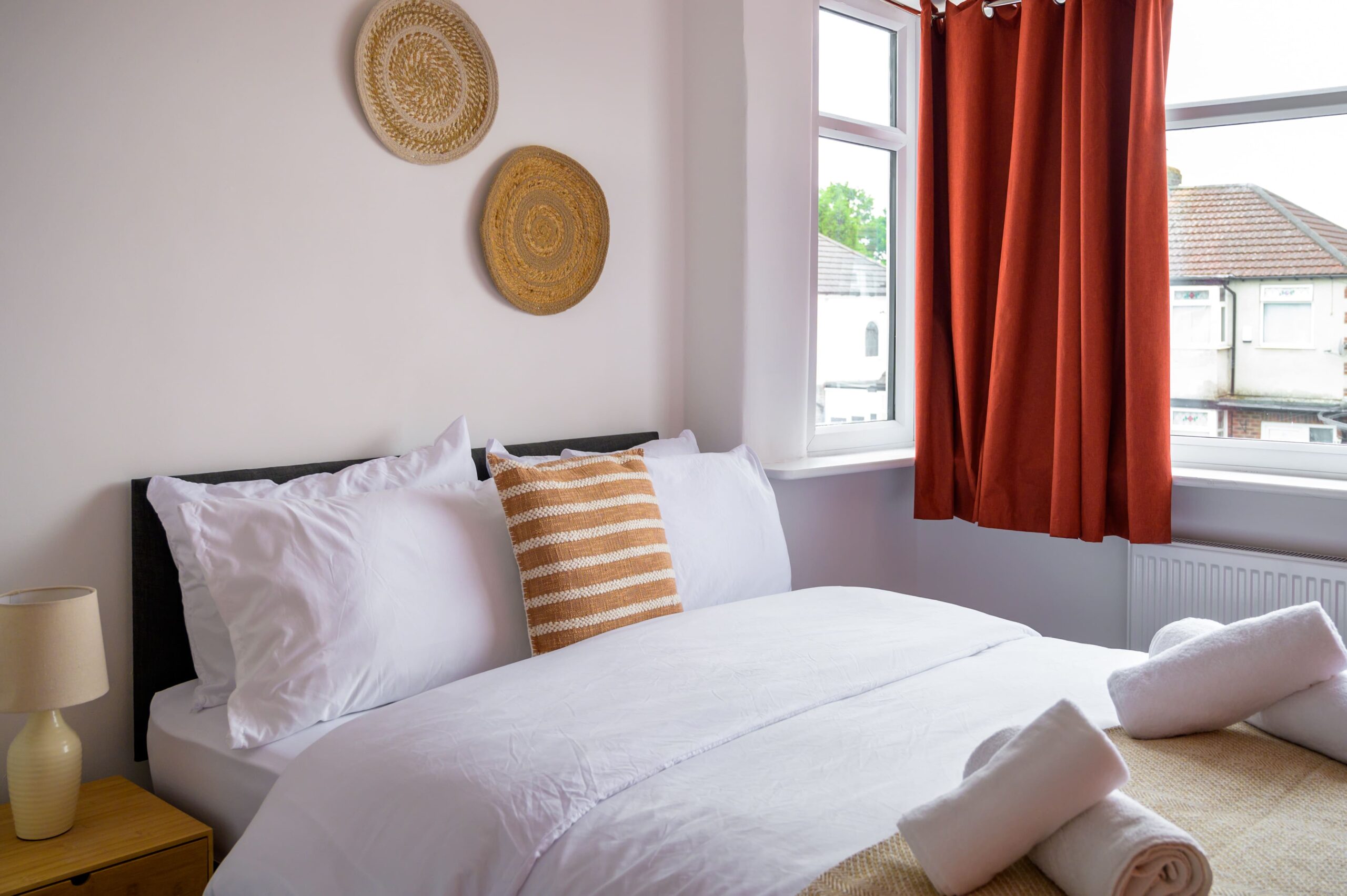 Bright bedroom with white bedding, an orange striped cushion, woven wall art, and orange curtains by a window