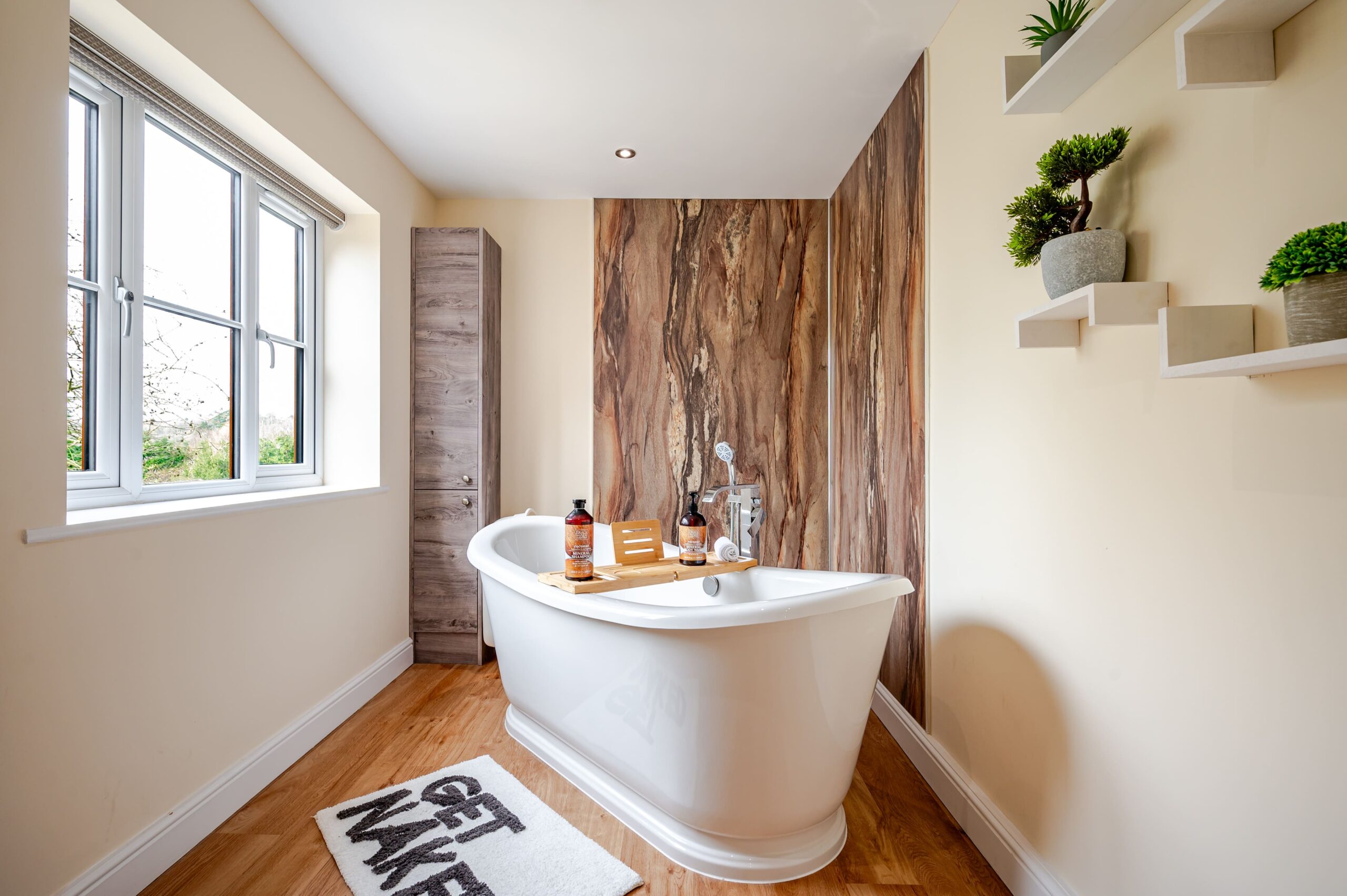 Modern bathroom featuring a white freestanding bath, wood-effect wall panel, tall cabinet, and wooden floors