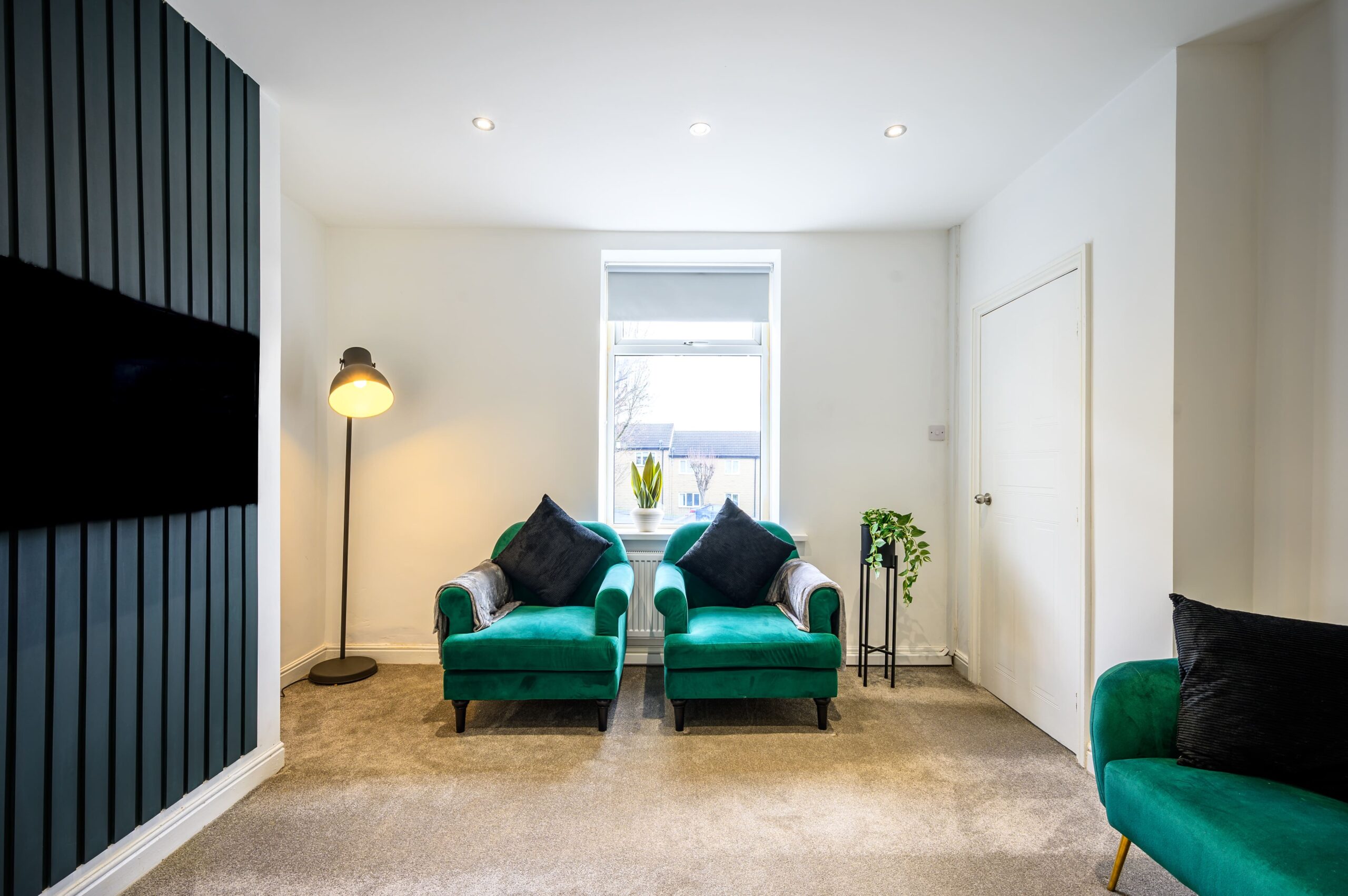 Two emerald green velvet armchairs in a modern living room with a floor lamp and dark slatted wall feature