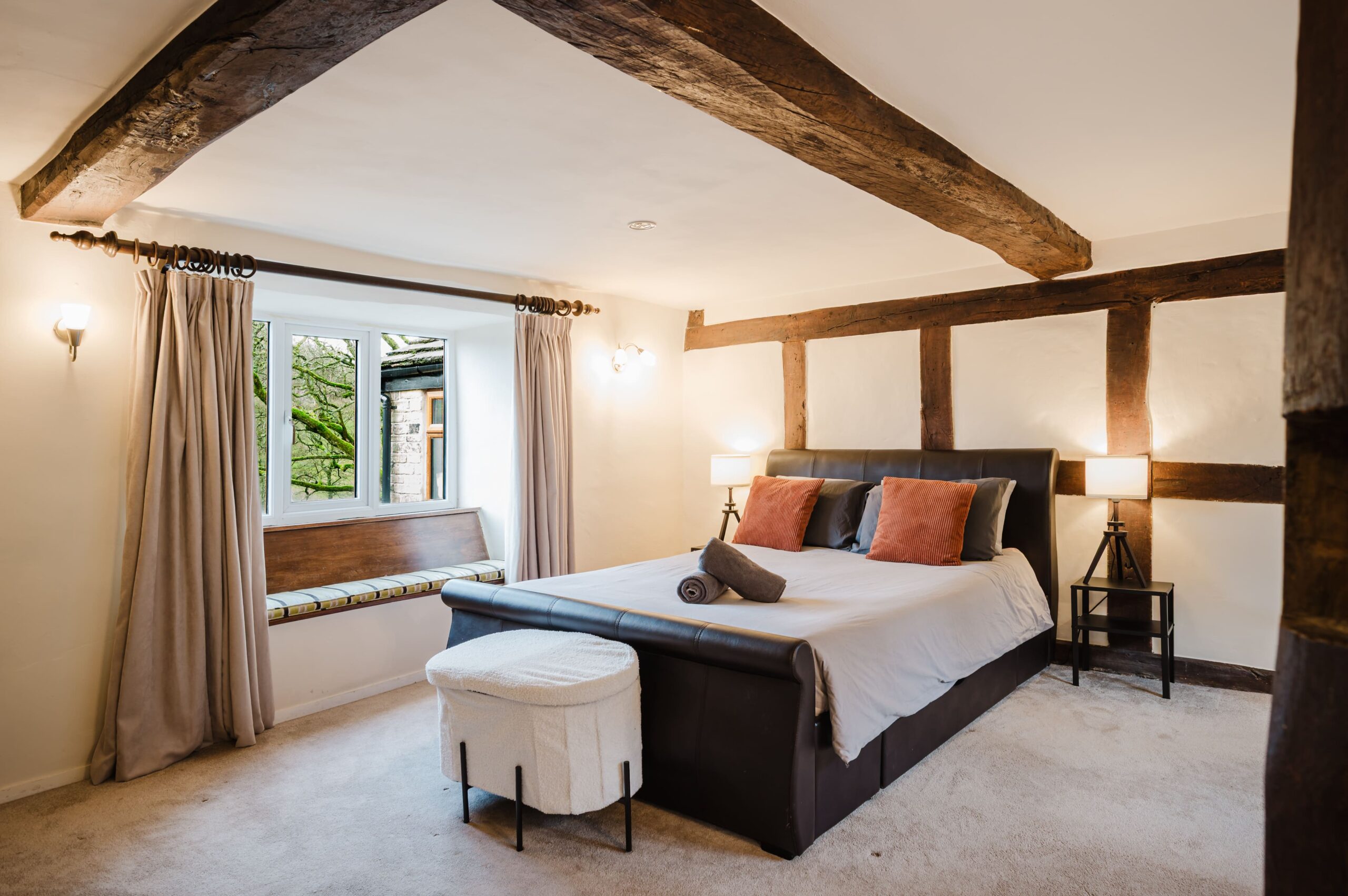 Bedroom with exposed wooden beams, a large bed with grey bedding and orange cushions, and a window seat