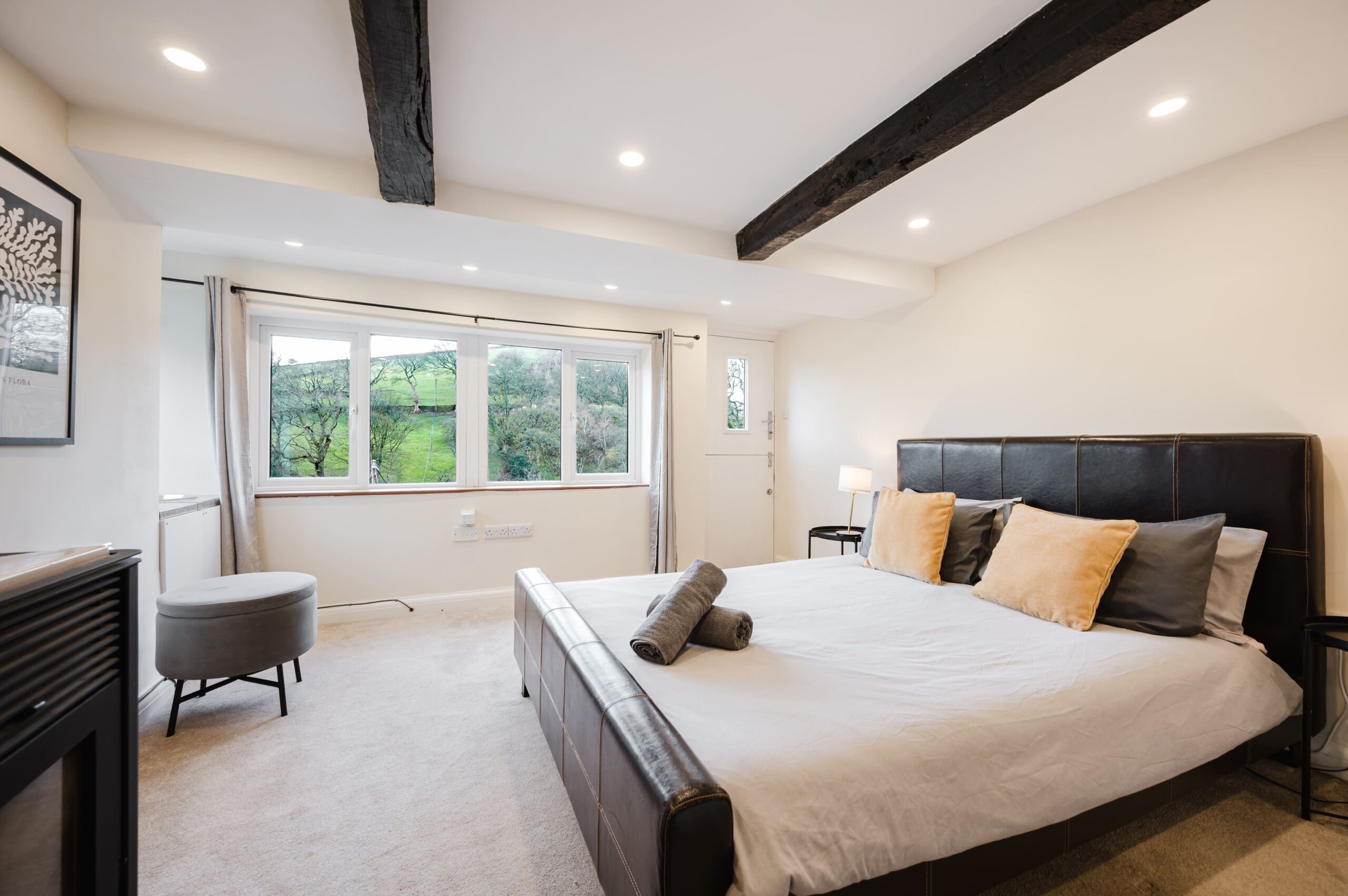 Modern bedroom with a leather bed, rustic ceiling beams, and a large window overlooking green hills