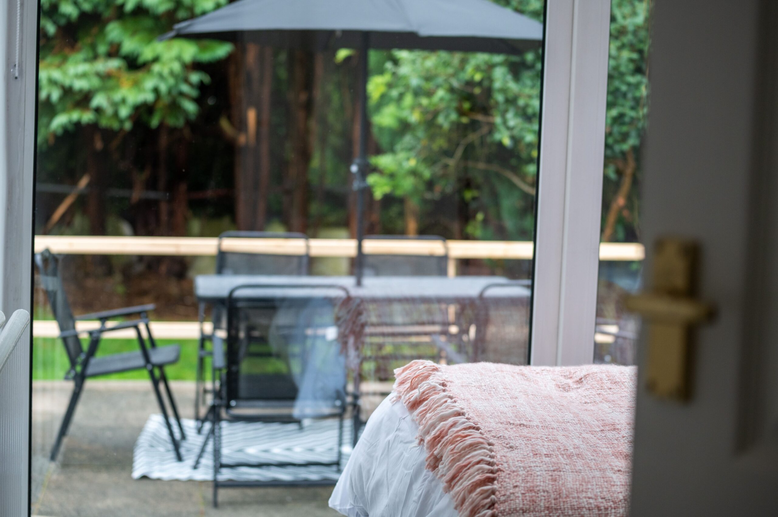 View from a bedroom looking out onto a patio with outdoor furniture and a parasol, surrounded by trees