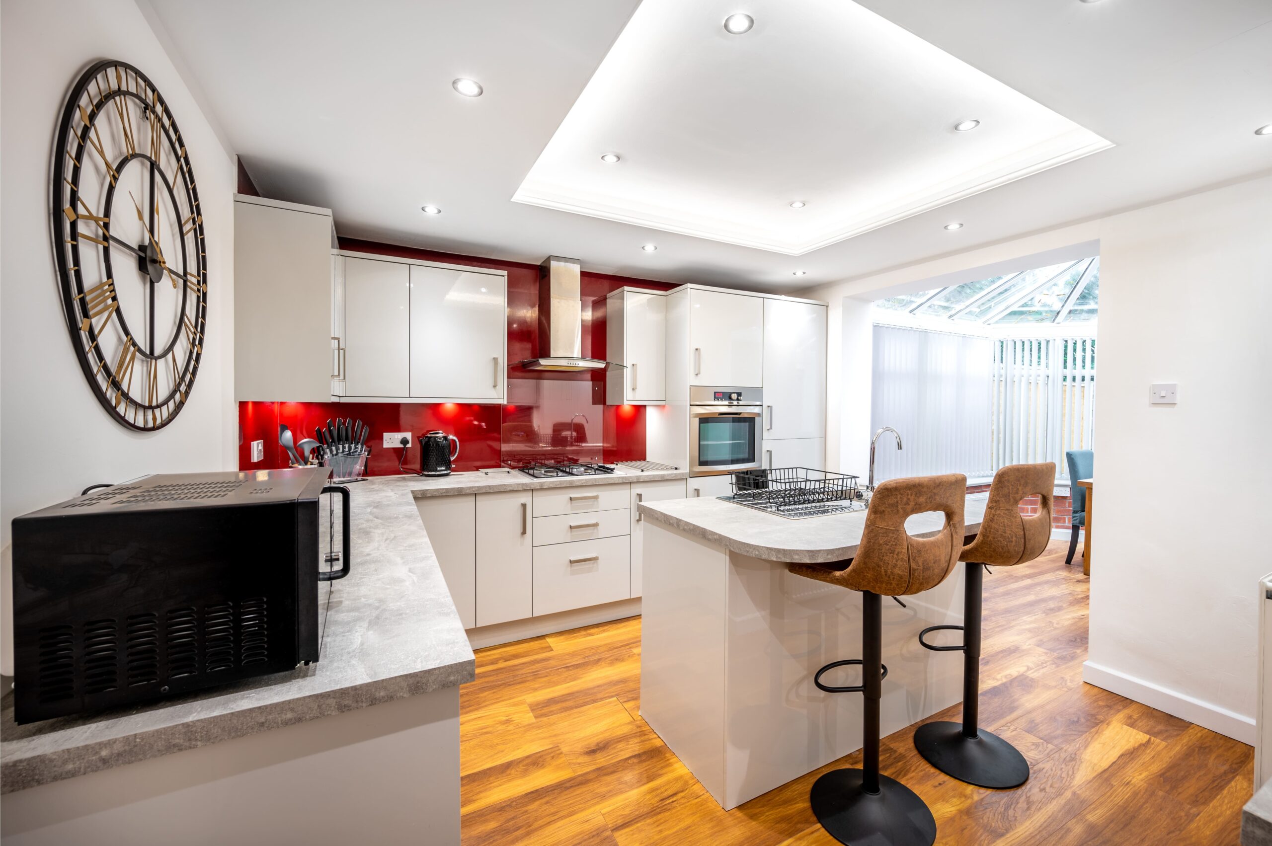Modern kitchen with white cabinets, red splashback, kitchen island with bar stools, and large wall clock