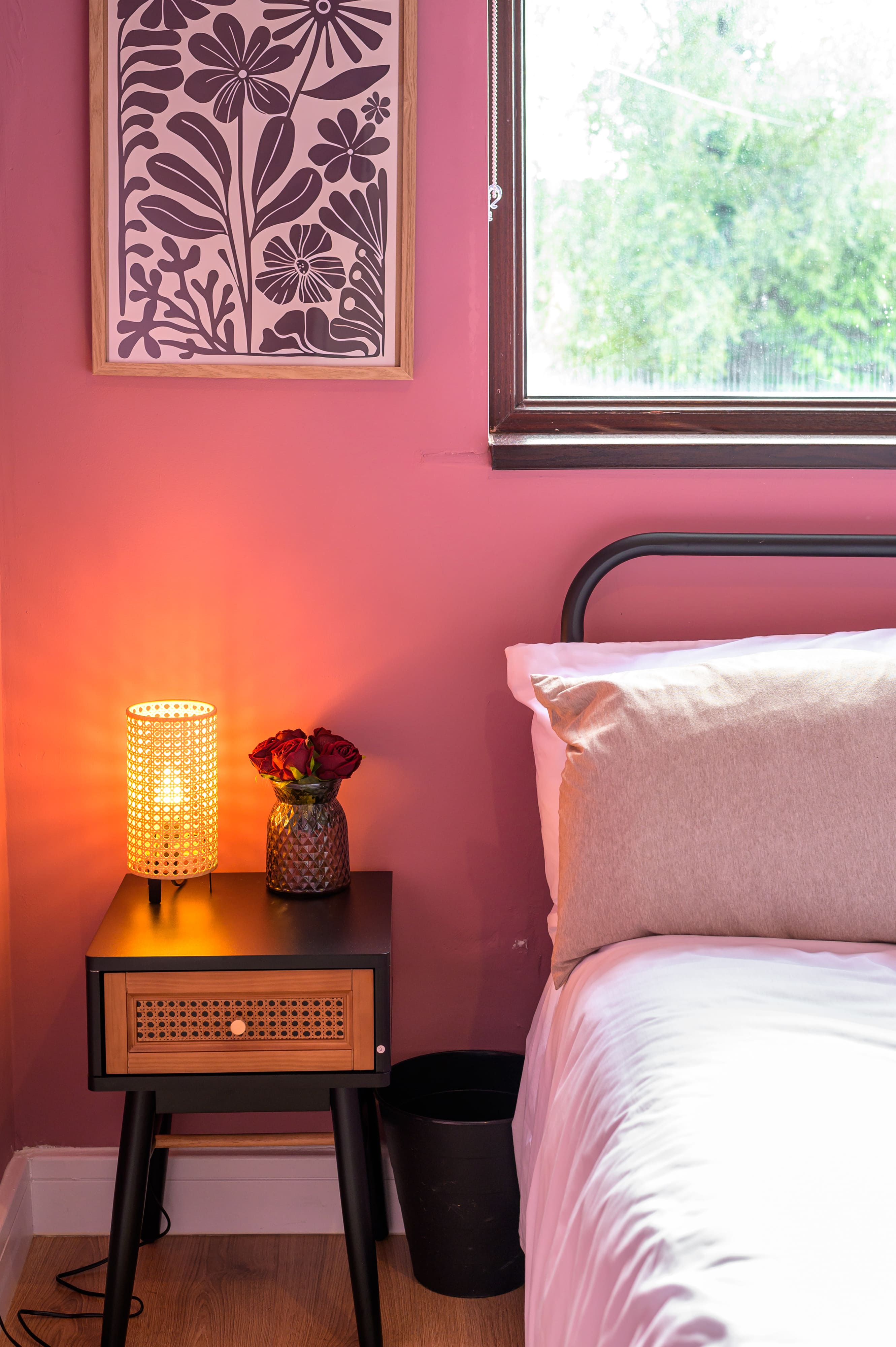 Cosy bedroom corner with pink walls, a glowing rattan lamp on a black bedside table, and a bed with white bedding