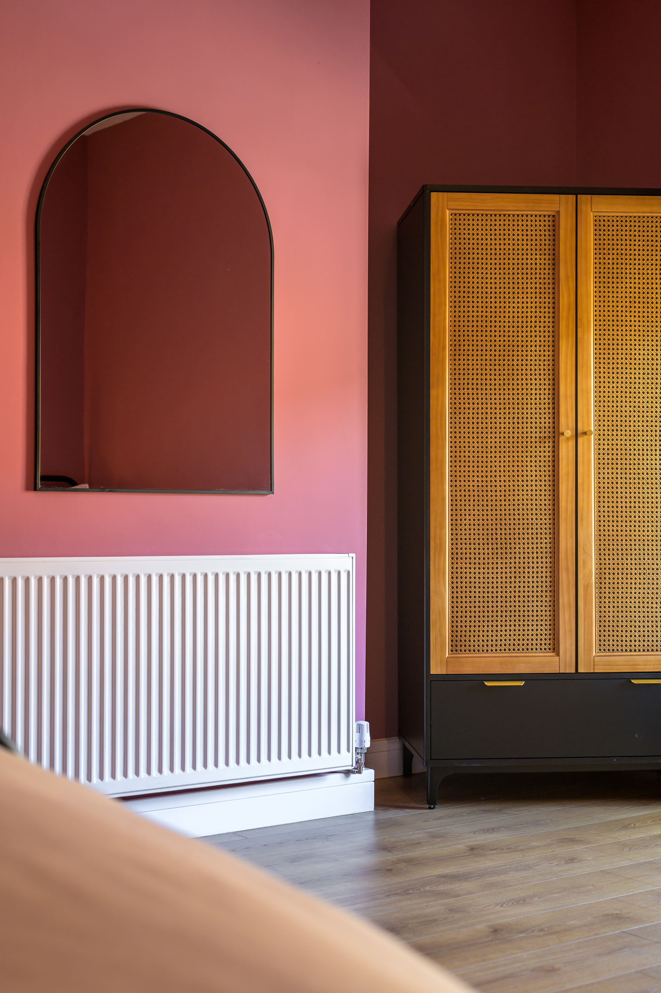 Pink wall featuring an arched mirror above a white radiator, beside a black wardrobe with rattan door panels