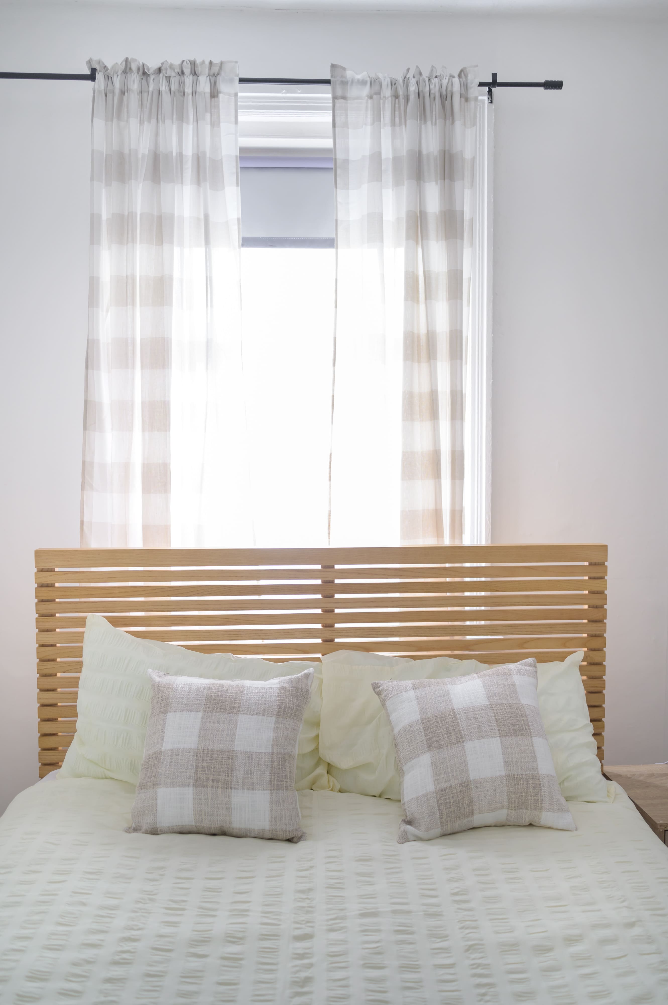Bed with wooden slatted headboard, cream bedding, and checked cushions in front of a window with sheer curtains