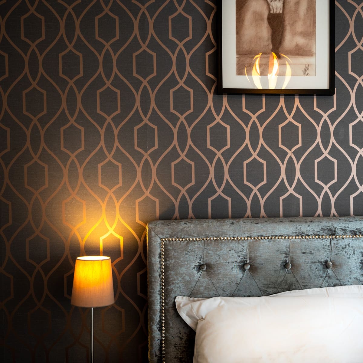 A bedroom with dark geometric wallpaper, a grey velvet tufted headboard, and a glowing bedside lamp
