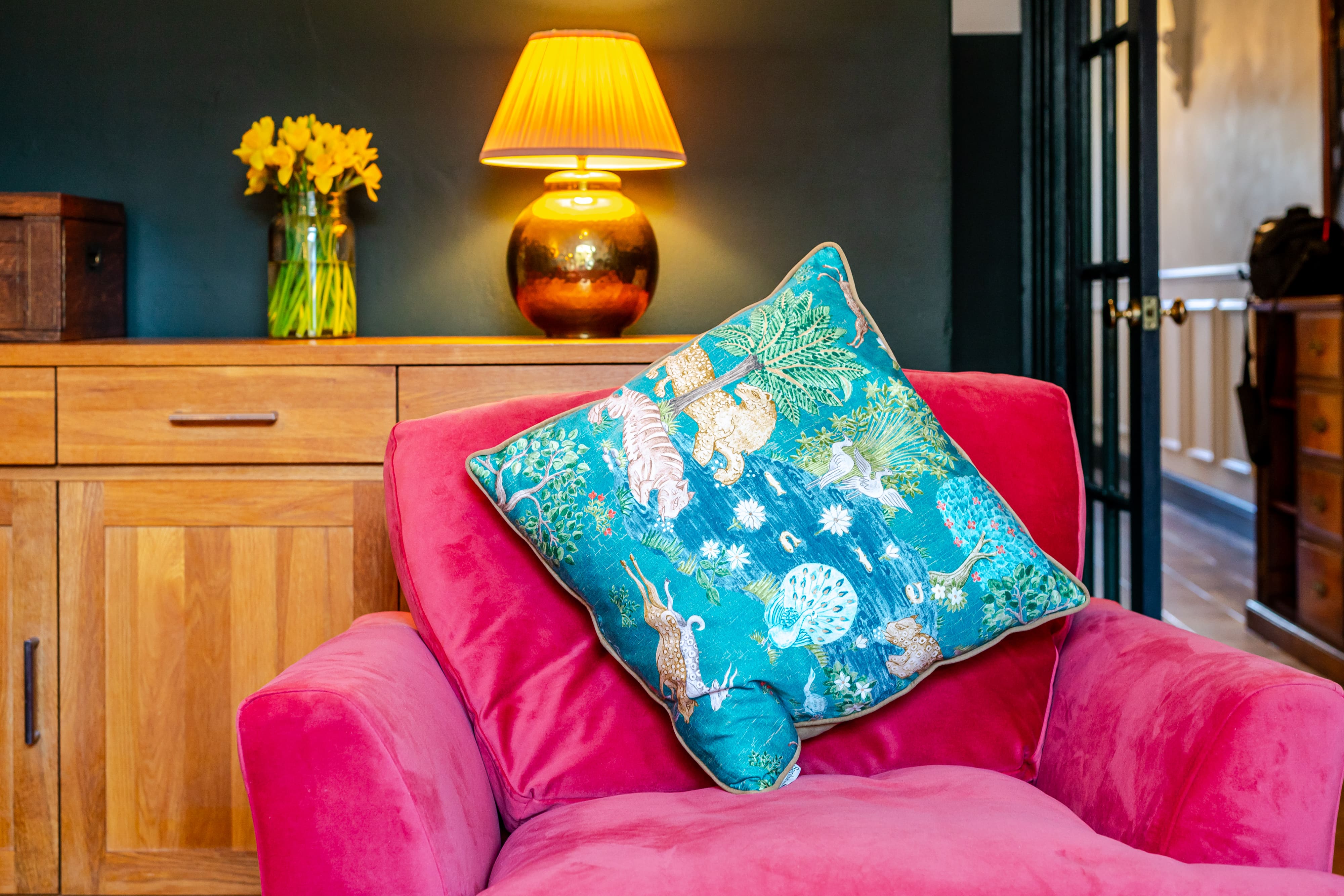 Pink velvet armchair with a teal animal-patterned cushion in a room with a lit lamp and yellow daffodils