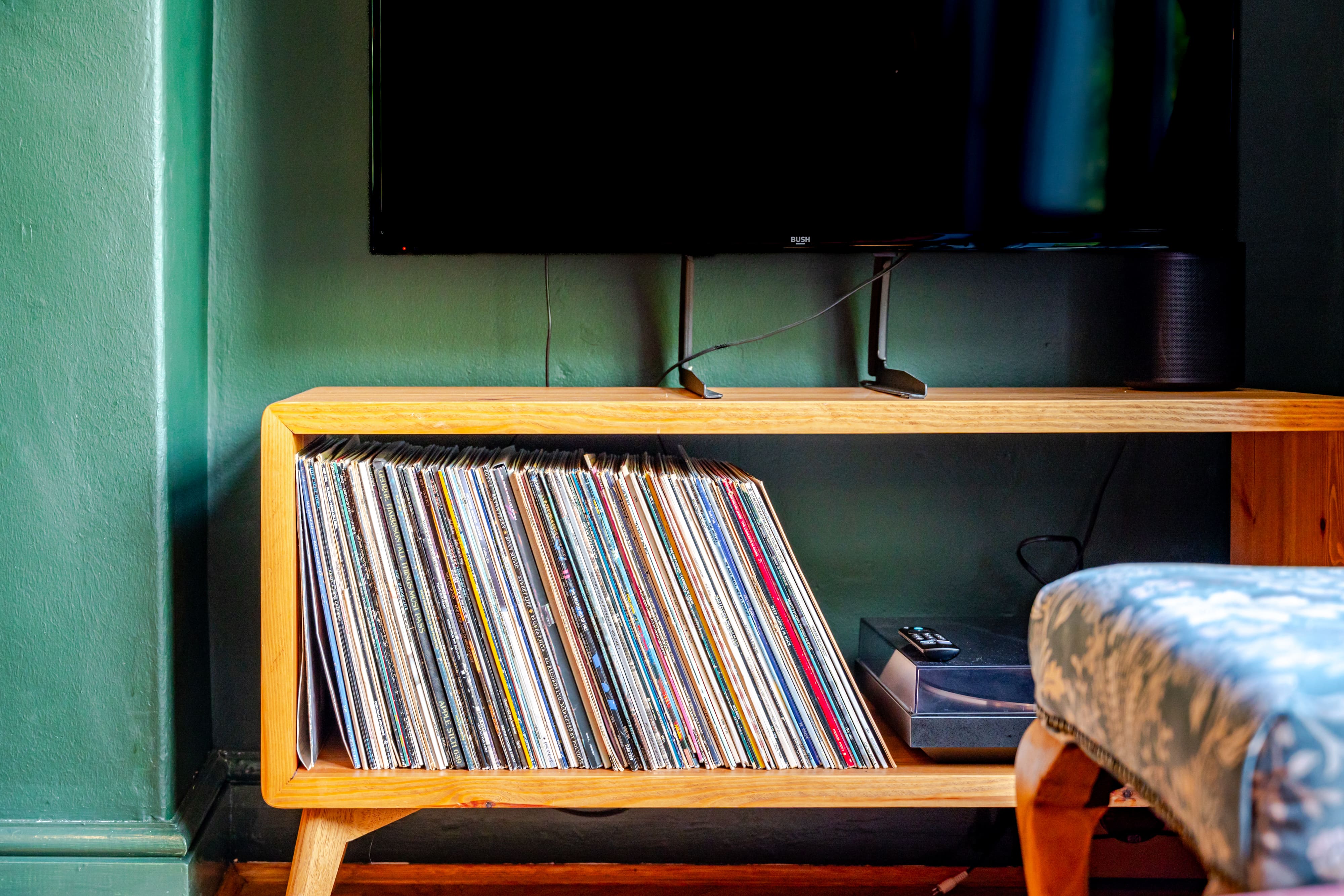Vinyl records stacked in a wooden media console beneath a television against a dark green wall