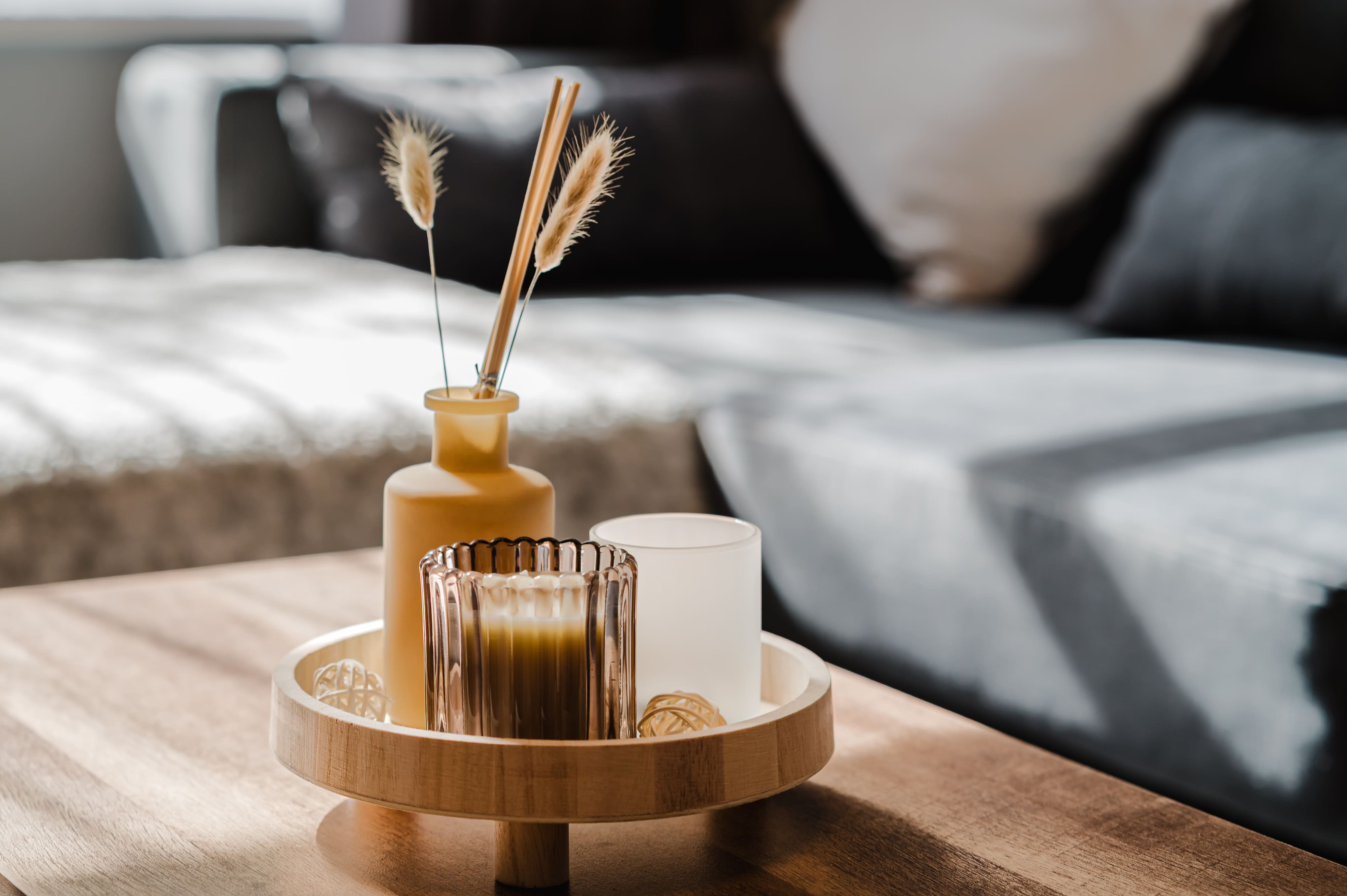 A wooden tray on a coffee table holding a yellow vase with dried grass and two candles in a sunlit living room