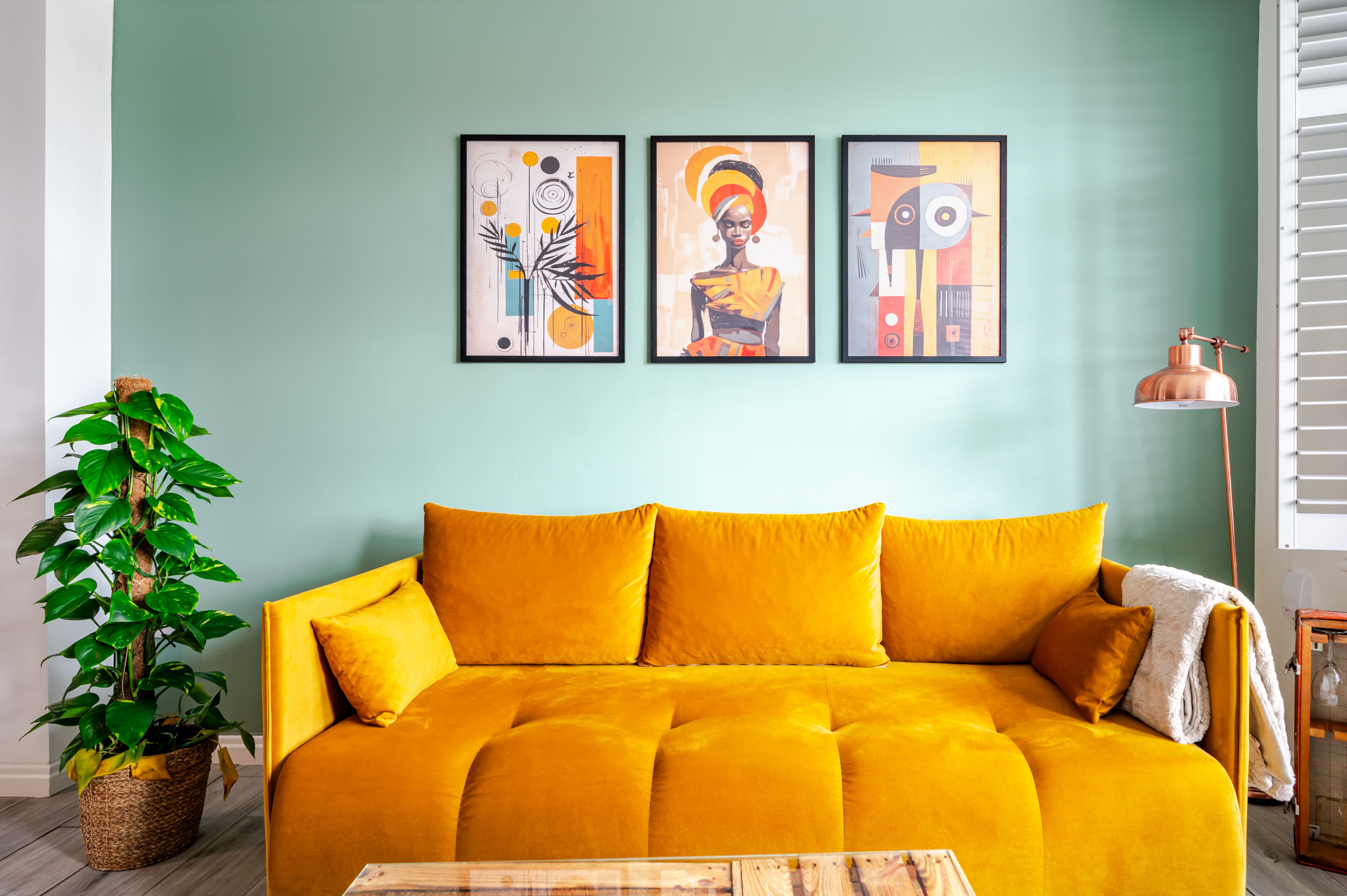 Bright yellow velvet sofa against a pale green wall with three framed art prints and a tall indoor plant