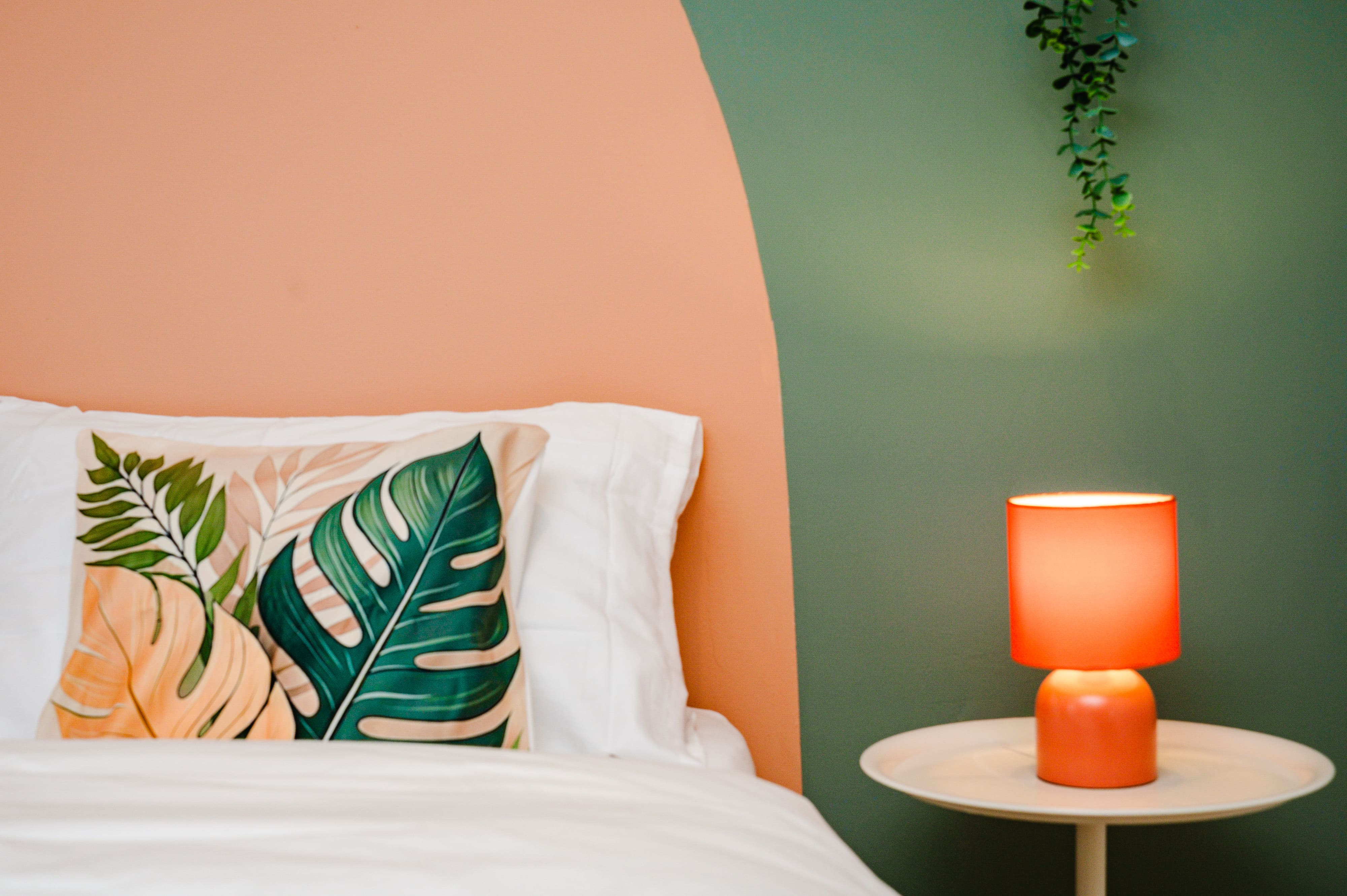 Bed with a tropical leaf pillow next to an orange lamp on a side table against a green and peach wall