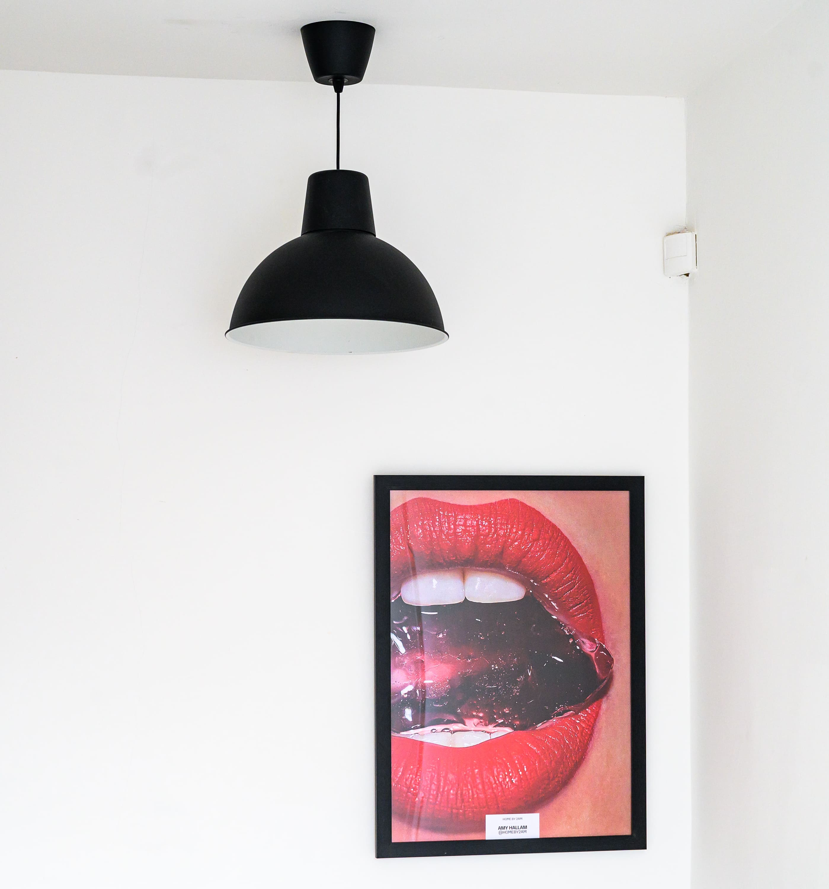 A black pendant light hanging above a framed art print of red lips on a white wall