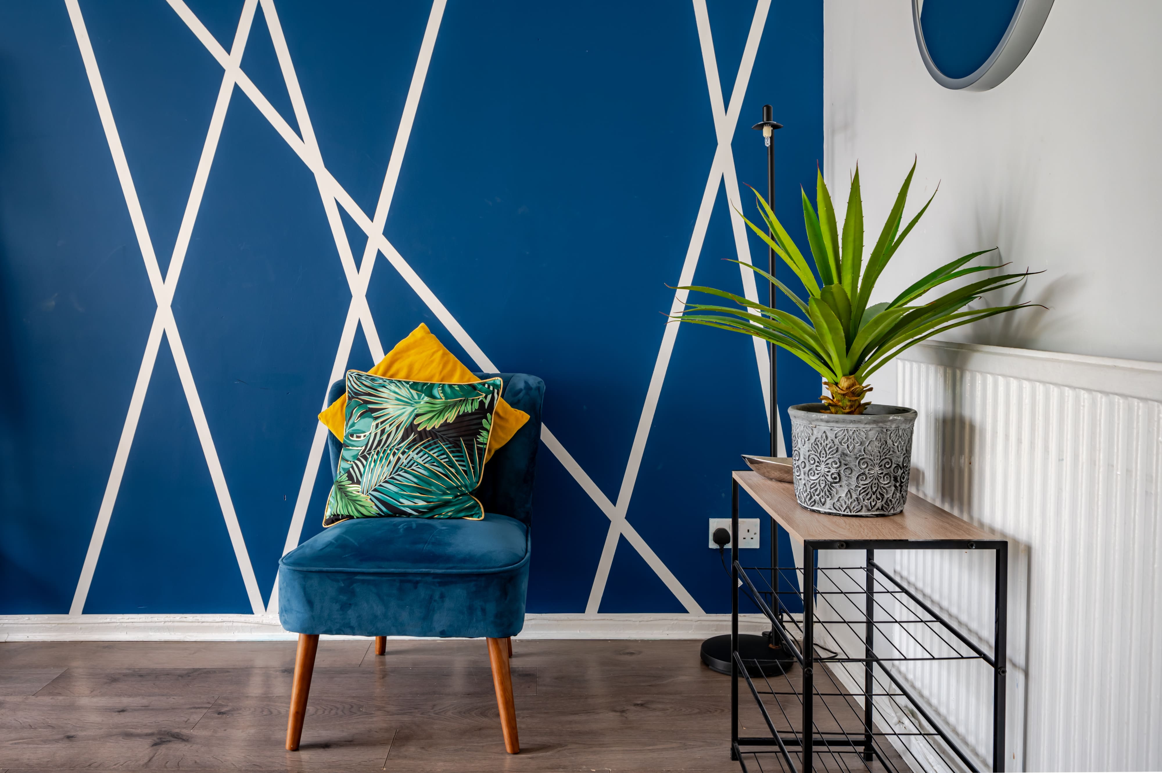 Blue velvet armchair with cushions against a blue wall with white geometric lines, next to a potted plant