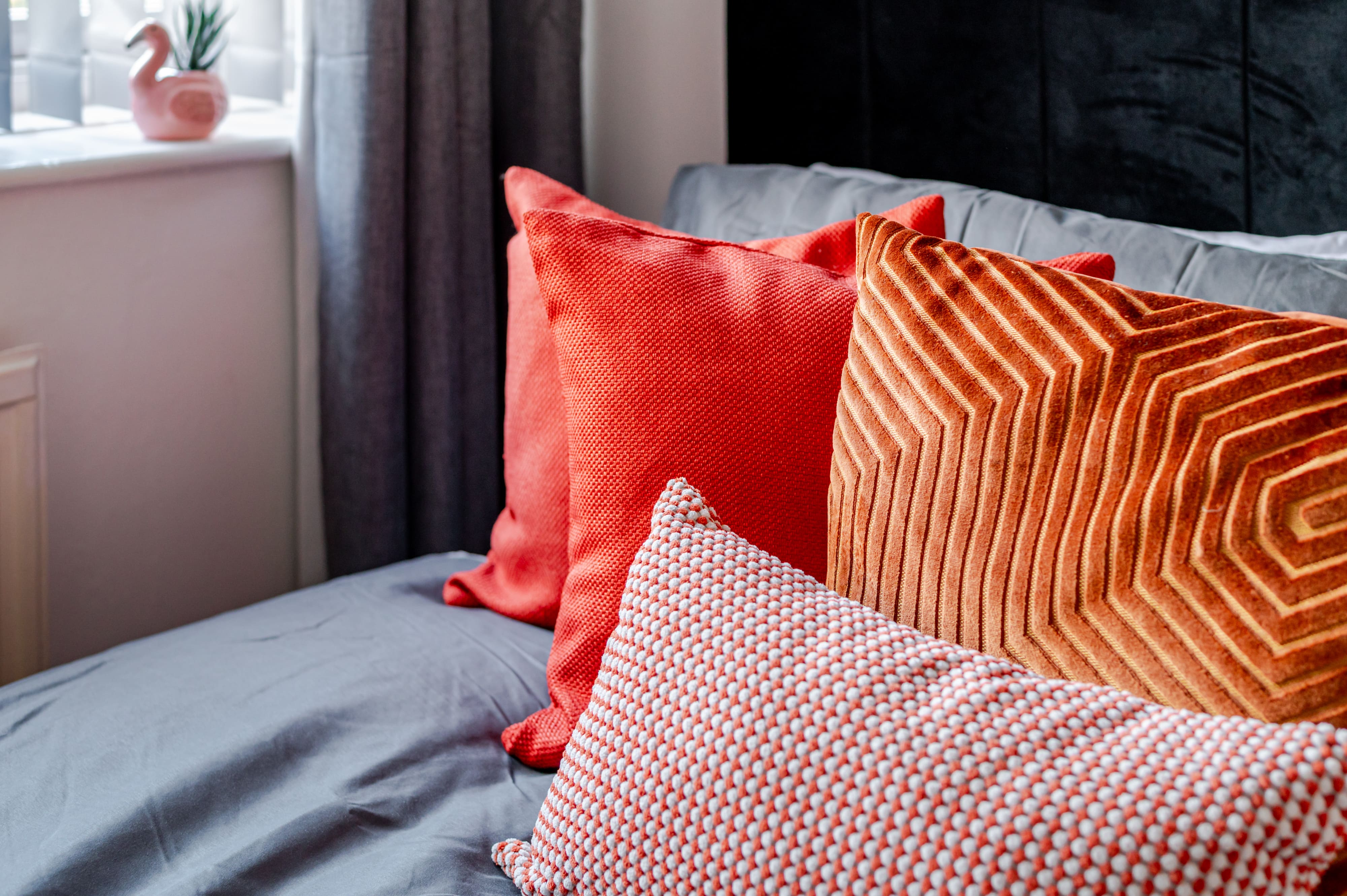 Orange and red decorative cushions with various textures and patterns arranged on a grey bed