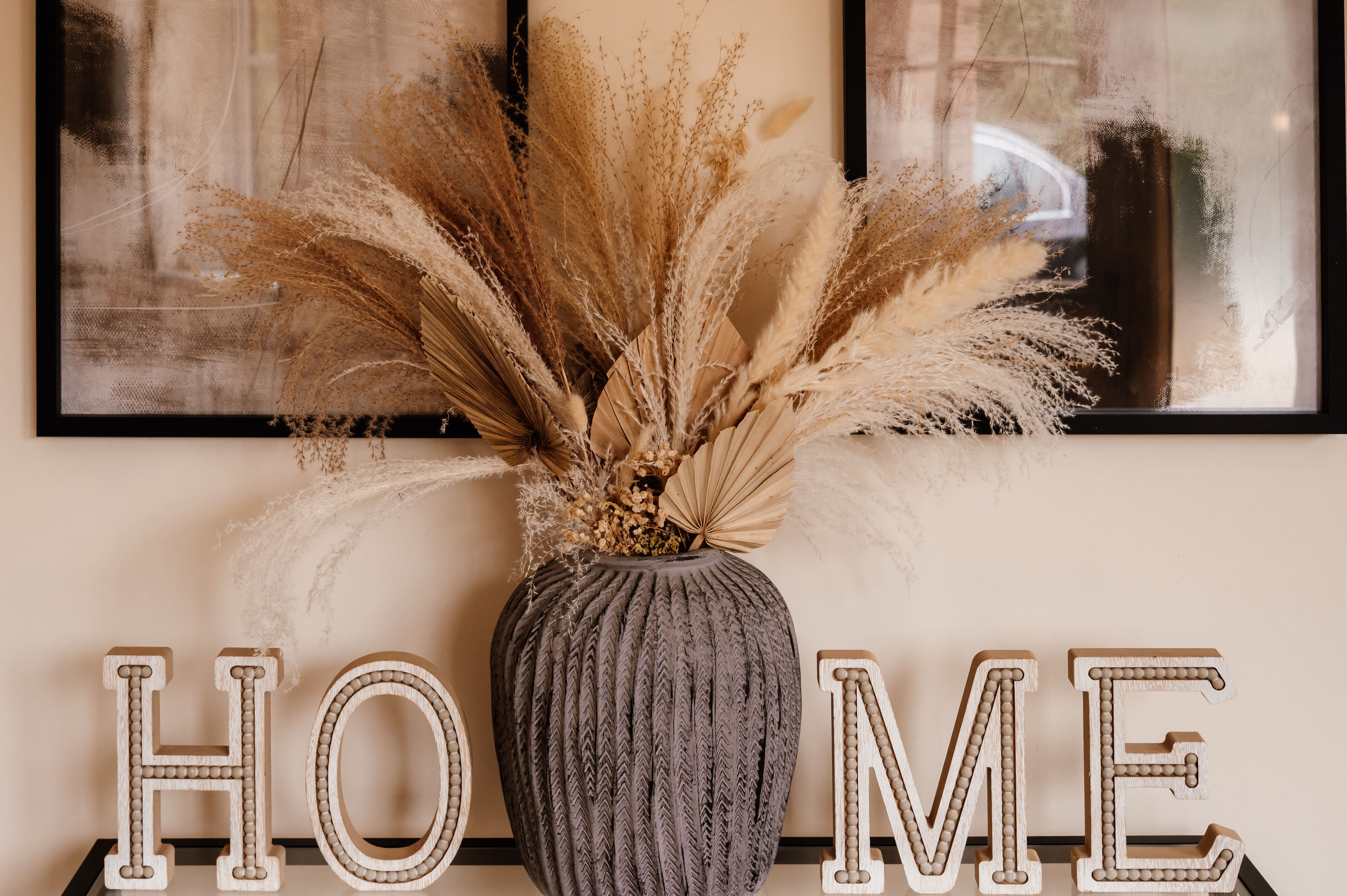Dried pampas grass in a grey vase between wooden letters spelling HOME, set against a wall with framed art